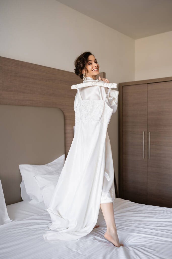 Ευτυχισμένη νεαρή νύφη με καστανά μαλλιά σε λευκό μεταξωτό χιτώνα κρατώντας μαλακή κρεμάστρα με κομψό νυφικό και στέκεται στο κρεβάτι στο υπνοδωμάτιο του δωματίου του ξενοδοχείου, ειδική περίσταση, όμορφη γυναίκα  - Φωτογραφία, εικόνα