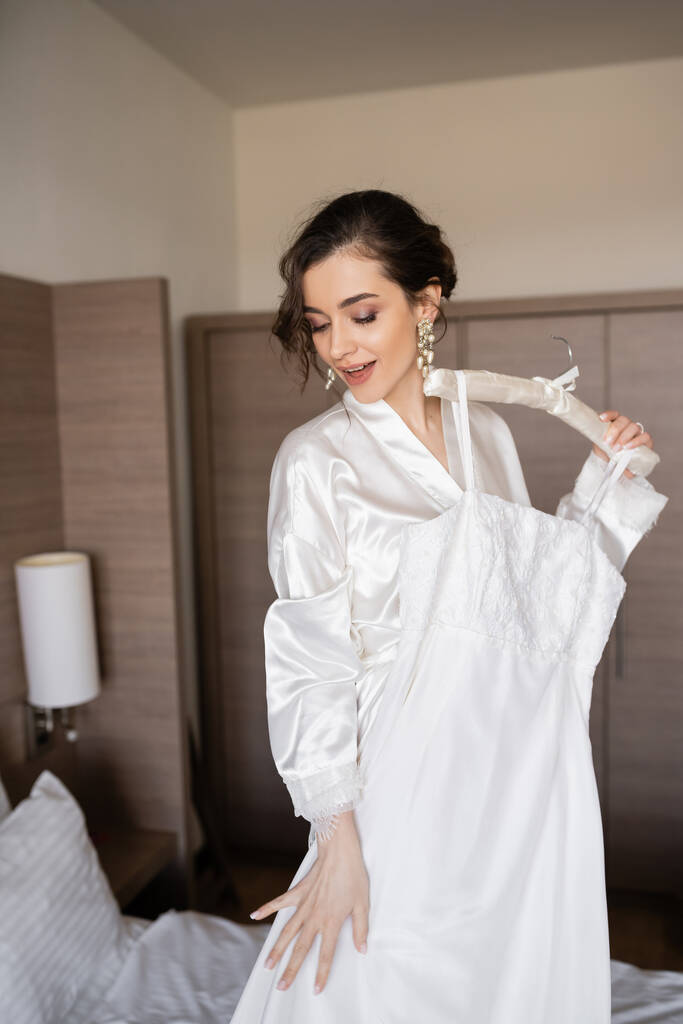 erstaunte junge Braut mit brünetten Haaren im weißen Seidenmantel mit weichem Kleiderbügel und elegantem Hochzeitskleid im Schlafzimmer des Hotelzimmers, besonderer Anlass  - Foto, Bild