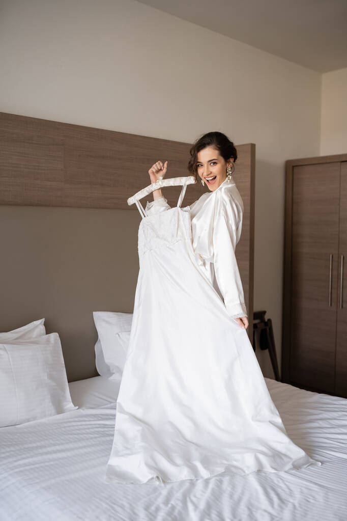 vzrušená nevěsta s brunetkou vlasy stojící v bílém hedvábí župan drží měkký věšák s elegantní svatební šaty a stojí na posteli v ložnici hotelového pokoje, zvláštní příležitosti, okouzlující žena  - Fotografie, Obrázek