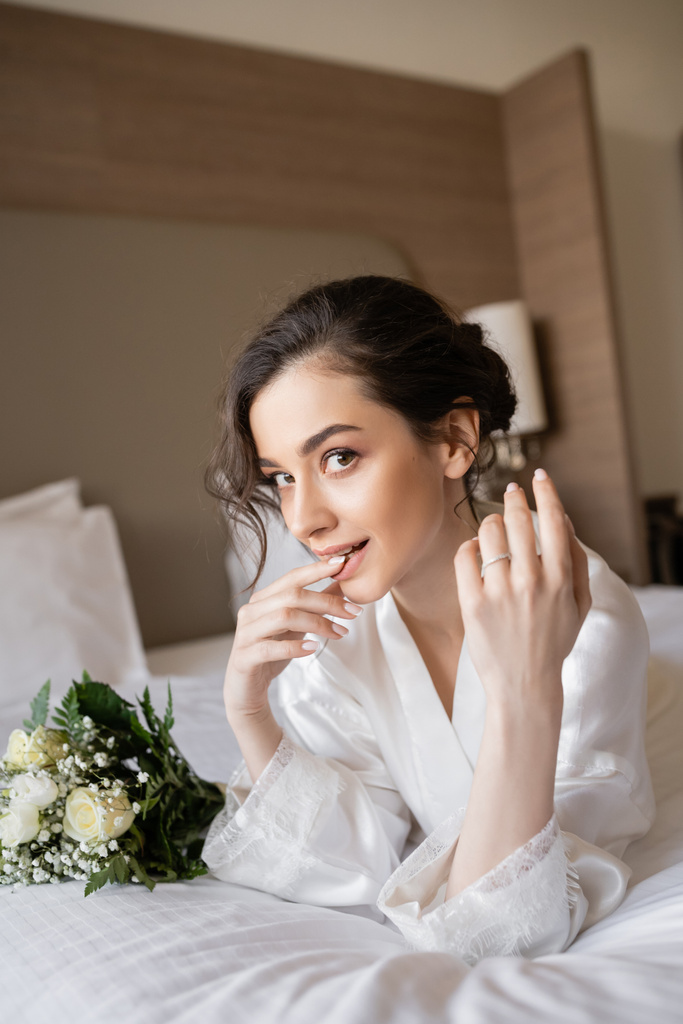 mulher encantadora com cabelo morena deitado em roupão de seda branca e mostrando anel de noivado no dedo ao lado do buquê de noiva no quarto do hotel no dia do casamento, ocasião especial, jovem noiva - Foto, Imagem