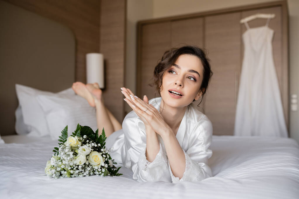 gorgeous woman with brunette hair and engagement ring on finger looking away and lying in white silk robe next to bridal bouquet on bed with wedding dress on blurred background, young bride - Photo, Image