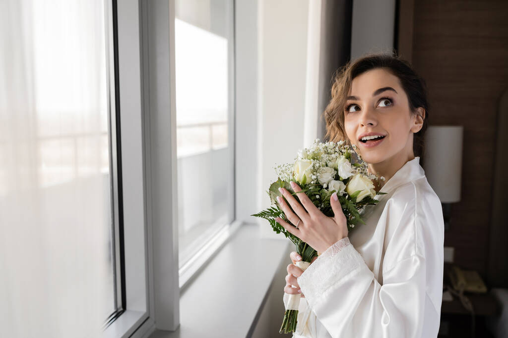 verbaasd jonge vrouw met verlovingsring om vinger staan in witte zijden mantel en houden bruids boeket terwijl kijken omhoog naast raam in hotel suite, speciale gelegenheid, bruid op trouwdag - Foto, afbeelding