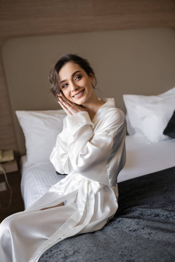 delightful and smiling bride with brunette hair sitting in white silk robe on comfortable bed with black blanket and looking away in hotel suite on wedding day, special occasion  - Photo, Image