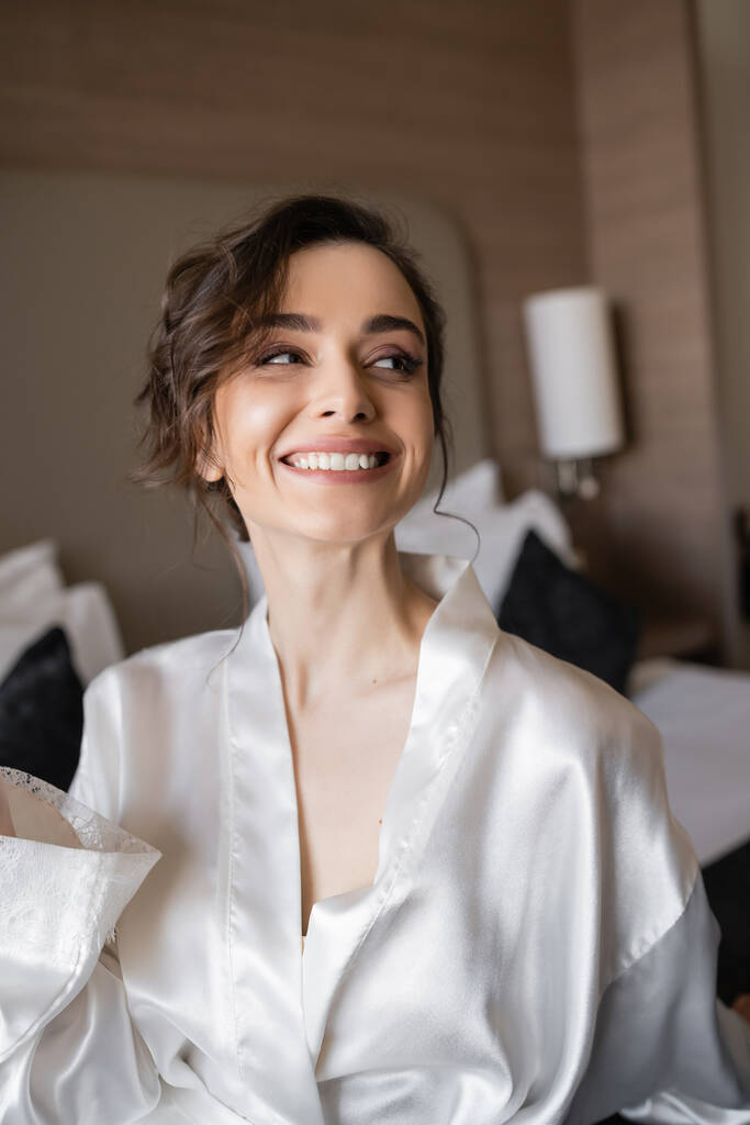 восхитительная женщина с брюнетками и свадебный макияж в белом шелковом халате улыбается и смотрит прочь в номере отеля в день свадьбы, особый случай  - Фото, изображение