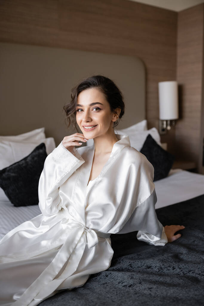 elégedett menyasszony barna hajjal és menyasszonyi smink megható áll és ül fehér selyem köntösben kényelmes ágyban, és nézi a kamera a hotel lakosztályában az esküvő napján  - Fotó, kép