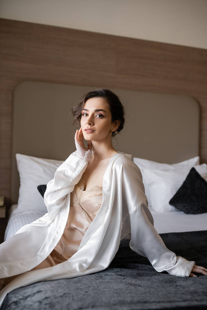 encantadora joven novia con cabello morena y maquillaje nupcial tocando su cara y sentado en bata de seda blanca en la cómoda cama de la suite del hotel el día de la boda  - Foto, imagen