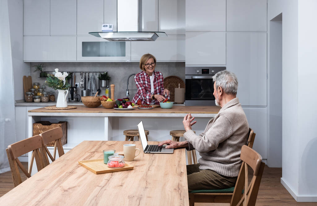 Happy senior volwassen man man afstandsbediening werken of afstand leren online vanaf kantoor met behulp van laptop computer, terwijl haar oudere vrouw koken ontbijt in de keuken. Familie van middelbare leeftijd thuis. - Foto, afbeelding