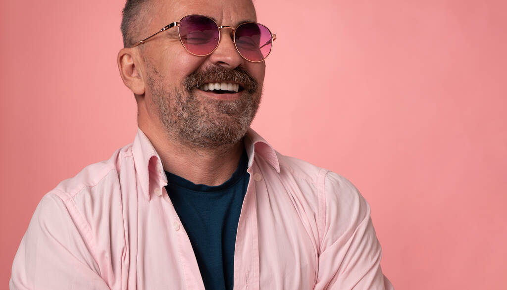 Portrét úspěšného pohledného usměvavého dospělého muže v růžových brýlích oblečeného v pastelově růžové košili izolované na růžovém pozadí. pohledný štětinatý muž tvář s velkým bílým úsměvem - Fotografie, Obrázek