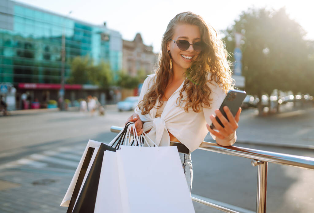 Szczęśliwa kobieta z torbami na zakupy spacerująca po słonecznej ulicy. Koncepcja stylu życia, kupowania, sprzedawania, konsumpcjonizmu. - Zdjęcie, obraz