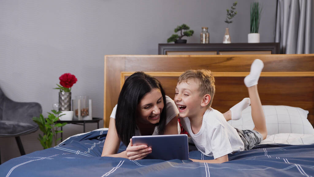 Charming high-spirited 30-aged brunette lying on comfortable bed with her handsome cheerful son and using tablet pc - Photo, Image