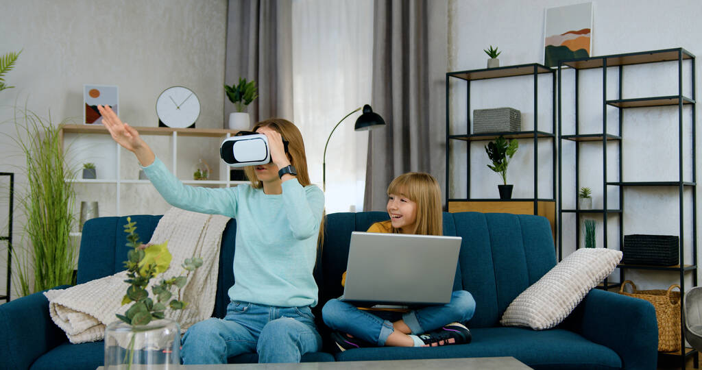 Hermoso positivo moderno dos diferentes edades niñas pasar ocio conjunto en jugar juegos virtuales con el ordenador portátil y gafas de realidad aumentada, vista frontal - Foto, imagen
