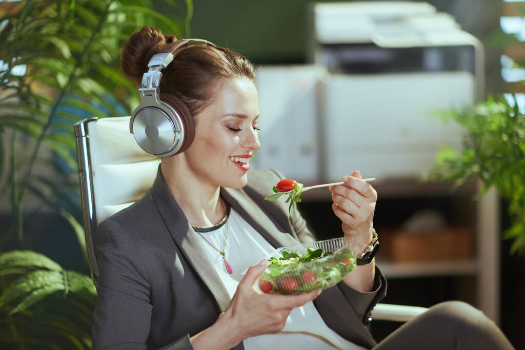 Un lieu de travail durable. femme propriétaire de petite entreprise moderne souriante dans un costume d'affaires gris dans un bureau vert moderne avec écouteurs manger de la salade. - Photo, image