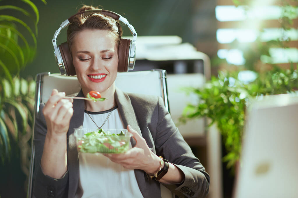 Sustainable workplace. smiling modern 40 years old bookkeeper woman in a grey business suit in modern green office with laptop eating salad and listening to the music with headphones. - Photo, Image