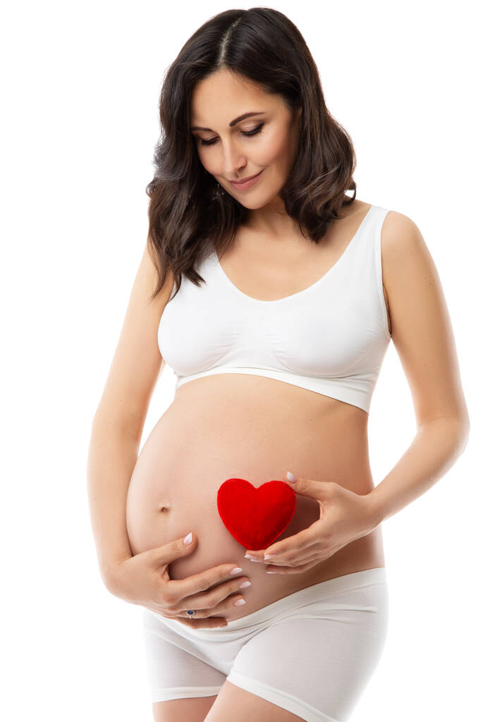Glückliche Schwangere mit rotem Herz am Bauch. Schwangerschaft Gesundheit Körperpflege. Schöne zukünftige Mutter in weißer Baumwollunterwäsche. Umstandspflege und Kosmetologie - Foto, Bild
