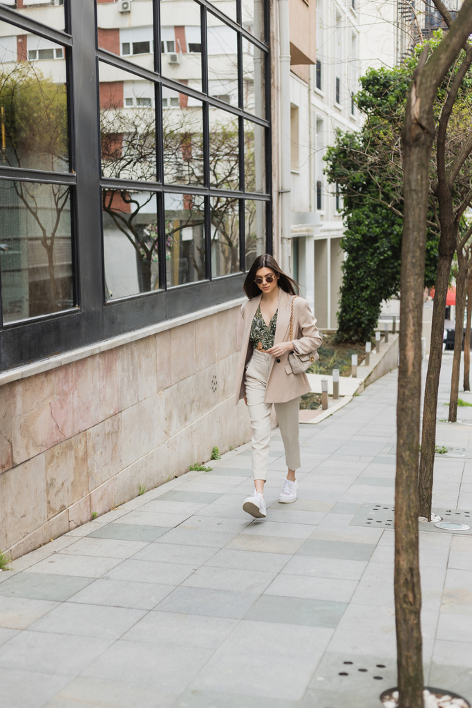 молодая брюнетка в стильных солнцезащитных очках и модном наряде с белыми брюками и бежевым блейзером прогулка с сумочкой рядом с деревьями и современное здание на улице в Стамбуле  - Фото, изображение