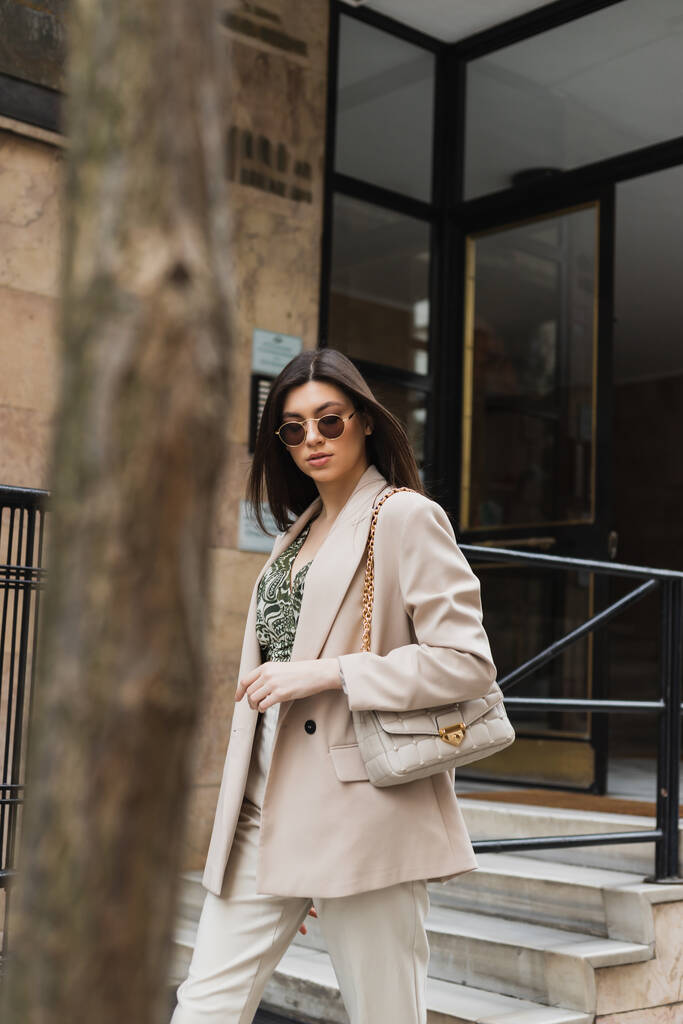 jeune femme brune avec des lunettes de soleil élégantes et une tenue tendance avec un pantalon blanc et un blazer beige marchant avec un sac à main près du bâtiment moderne et un tronc d'arbre flou dans la rue à Istanbul  - Photo, image