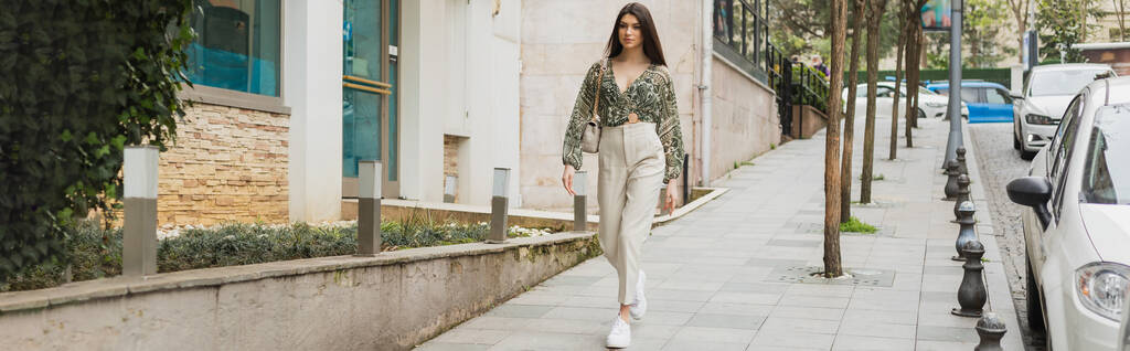mladá žena s dlouhými vlasy v béžových kalhotách, střiženou blůzou a kabelkou s řetízkovým řemínkem procházka v blízkosti moderní budovy, auta a zelené stromy na městské ulici v Istanbulu, turista, prapor  - Fotografie, Obrázek