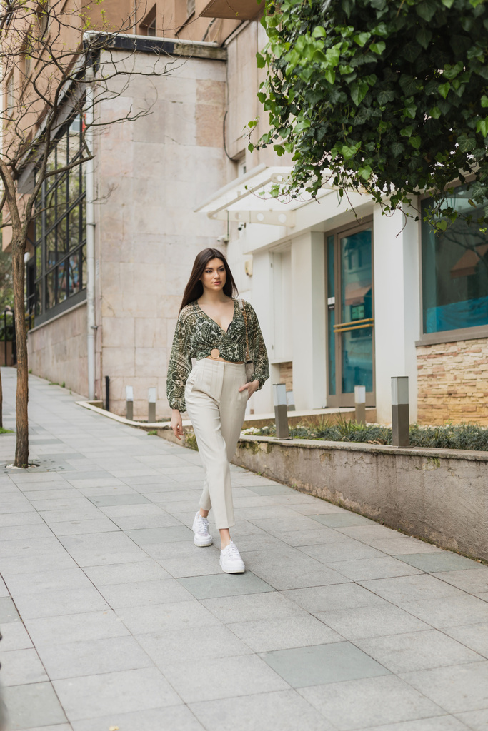 молода жінка з довгим волоссям в модному одязі з бежевими штанами, затиснутою блузкою і сумочкою з ланцюговим ременем, що йде з рукою в кишені біля сучасного будинку і зеленого дерева на вулиці в Стамбулі.  - Фото, зображення