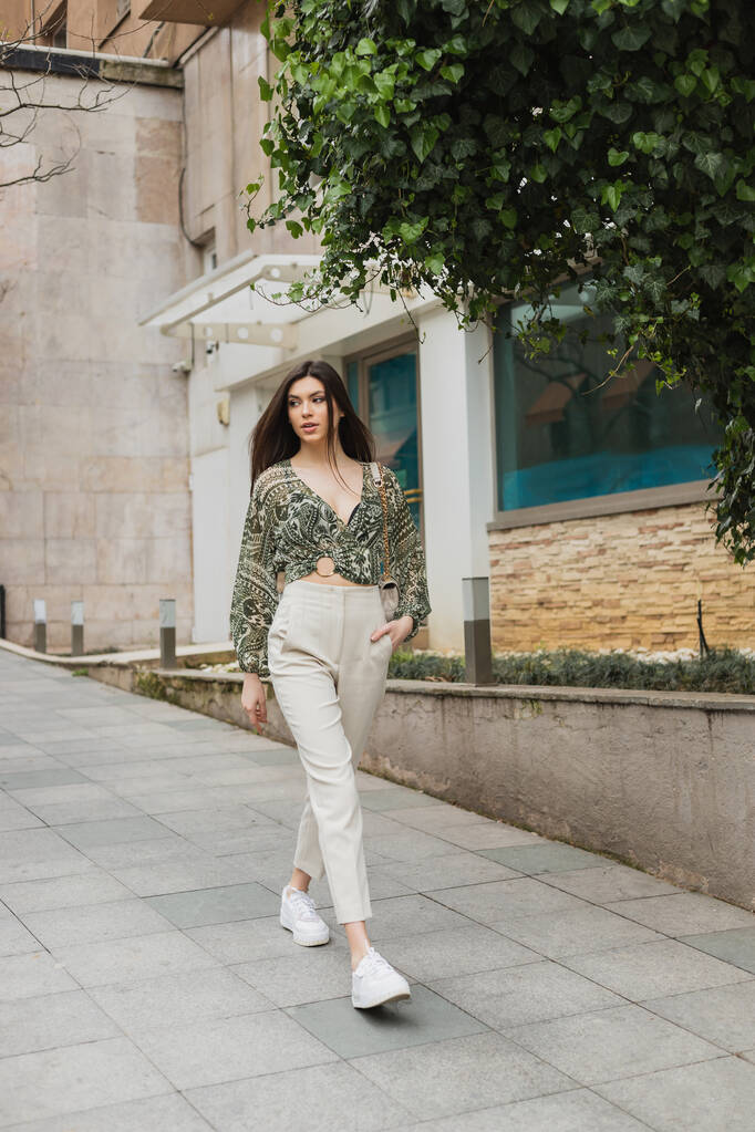 Uzun saçlı, modaya uygun giyinen, bej pantolonlu, kırpık bluzlu, kayışlı, modern binanın yanında el ele yürüyen, İstanbul caddesinde yeşil ağaçlı şık bir kadın.  - Fotoğraf, Görsel