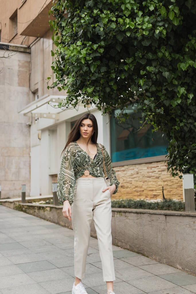 elegancka kobieta z długimi włosami w modnym stroju z beżowymi spodniami, przyciętą bluzką i torebką z paskiem na łańcuszku spacerującym z ręką w kieszeni w pobliżu nowoczesnego budynku i zielonym drzewem na ulicy miejskiej w Stambule  - Zdjęcie, obraz