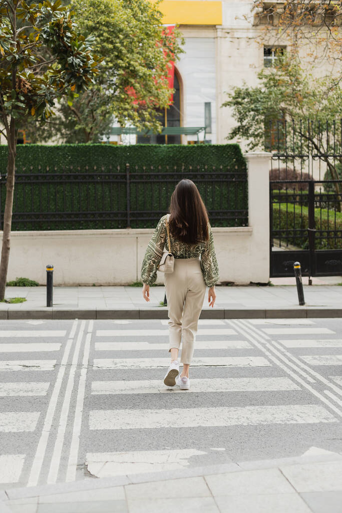 Rückansicht einer schicken Frau mit langen Haaren im trendigen Outfit mit beiger Hose, abgeschnittener Bluse und Handtasche mit Kettenriemen, die auf einem Fußgängerüberweg in Istanbul läuft - Foto, Bild