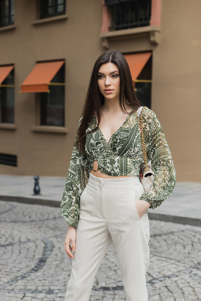 young woman with brunette long hair in beige pants, cropped blouse and handbag with chain strap standing with hand in pocket near blurred building on urban street in Istanbul  - Photo, Image