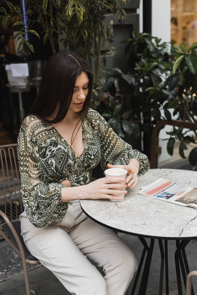νεαρή γυναίκα με μακριά καστανά μαλλιά και μακιγιάζ κάθεται στην καρέκλα κοντά στρογγυλό τραπέζι μπιστρό με εφημερίδα και κρατώντας χάρτινο κύπελλο με καφέ κοντά σε θολή φυτά στη βεράντα του καφέ στην Κωνσταντινούπολη  - Φωτογραφία, εικόνα