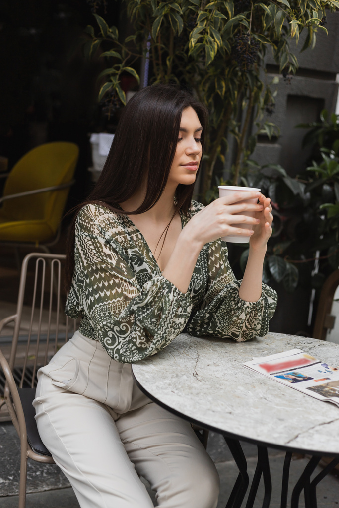長いブルネットの髪とメイクを持つ女性を魅了します新聞と丸いビストロテーブルの近くの椅子に座っているとイスタンブールのカフェのテラスでぼやけた植物の近くにコーヒーと紙コップを保持  - 写真・画像