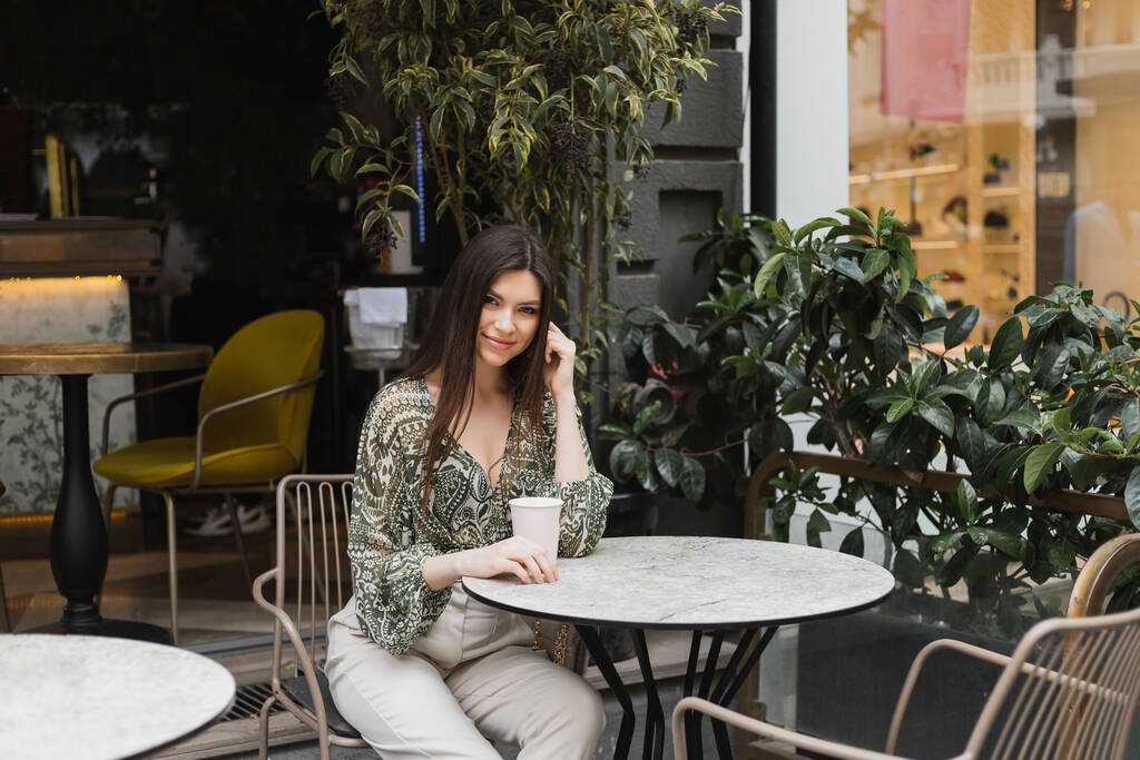 χαμογελαστή γυναίκα με μακριά καστανά μαλλιά και μακιγιάζ κάθεται στην καρέκλα κοντά στρογγυλό τραπέζι μπιστρό και κρατώντας καφέ σε χάρτινο κύπελλο κοντά θολή φυτά στη βεράντα του καφέ στην Κωνσταντινούπολη  - Φωτογραφία, εικόνα