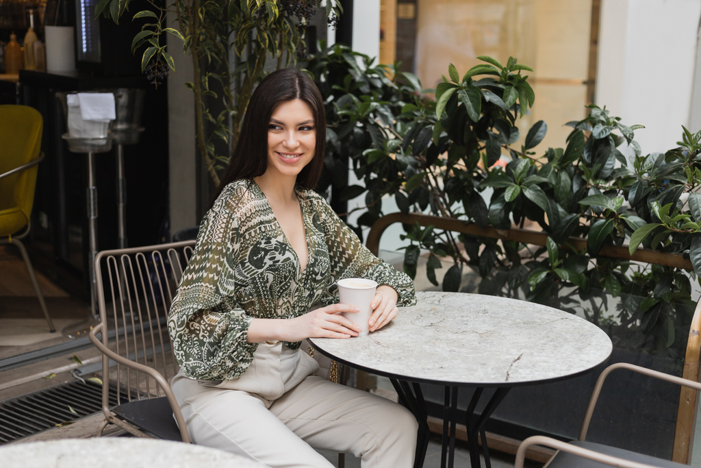 encantadora jovem com longos cabelos morena sentada na cadeira perto de mesa redonda bistrô e segurando café em xícara de papel enquanto olha para longe e sorrindo perto de plantas borradas no terraço do café em Istambul  - Foto, Imagem