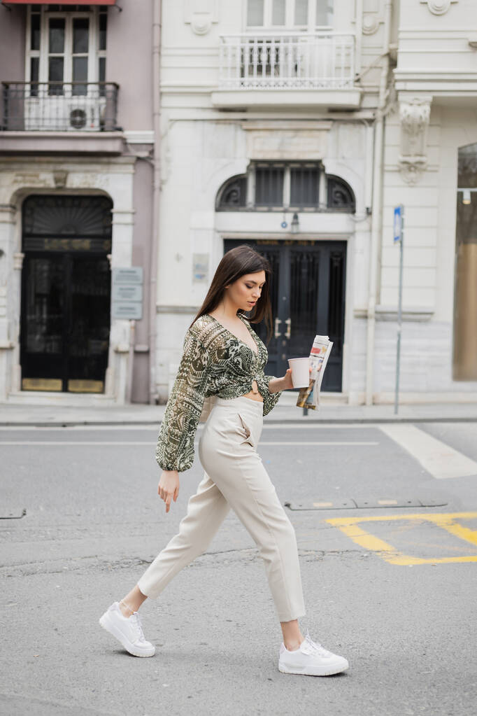 長いブルネットの髪とメイクを持つかなり若い女性イスタンブールの建物の近くの都市通りにベージュのパンツやブラウスと流行の衣装で歩いている間、コーヒーや新聞と紙カップを保持  - 写真・画像