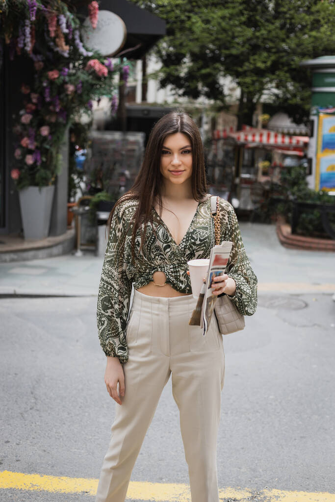 krásná žena s dlouhými vlasy drží papírový šálek s kávou a novinami, zatímco stojí v módní oblečení s kabelkou na řetízku popruh a usmívá se na městské ulici v blízkosti rozmazané květinářství v Istanbulu  - Fotografie, Obrázek
