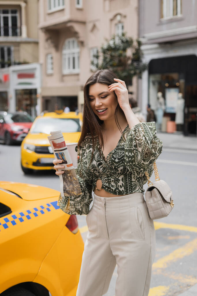 mulher feliz ajustando seu cabelo longo e segurando xícara de papel com café e jornal enquanto estava de pé na roupa da moda com bolsa na cadeia cinta perto de táxi amarelo na rua urbana turva em Istambul  - Foto, Imagem