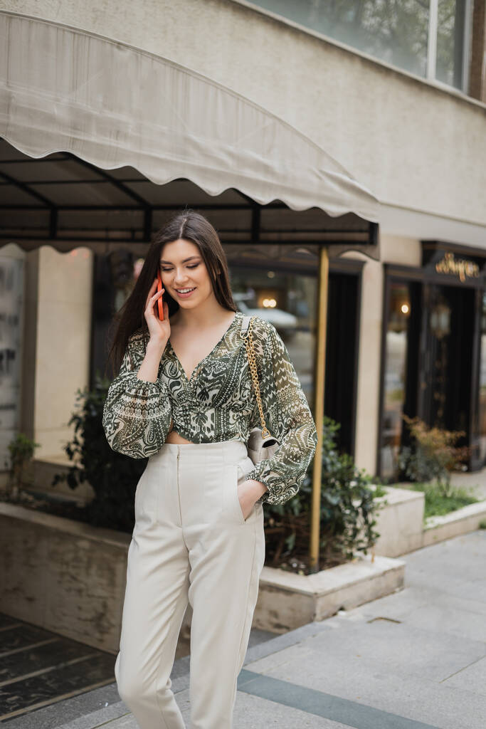 stylish young woman with brunette long hair and makeup smiling while talking on smartphone and standing in trendy outfit with handbag on chain strap near blurred fancy restaurant in Istanbul  - Photo, Image