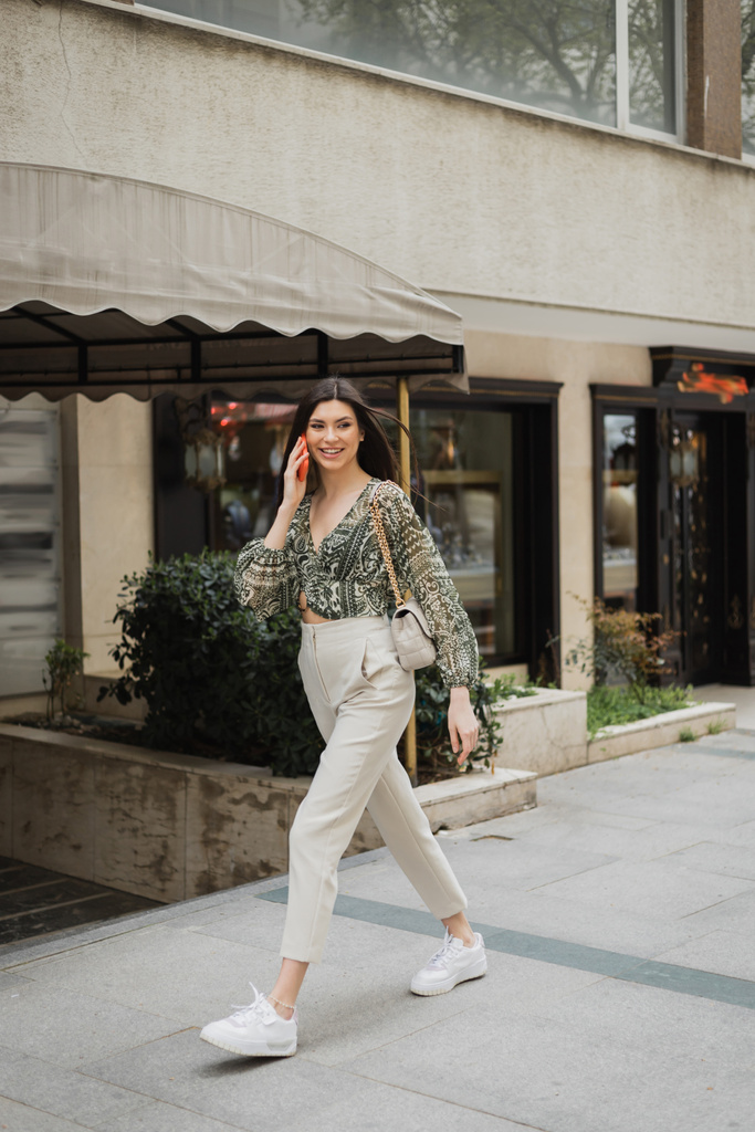 veselá mladá žena s brunetkou dlouhé vlasy a make-up s úsměvem při hovoru na smartphone a chůzi v módní oblečení s kabelkou na řetízku popruh v blízkosti rozmazané luxusní restaurace v Istanbulu  - Fotografie, Obrázek