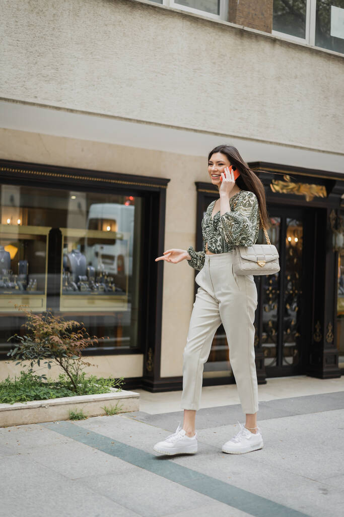 Uzun saçlı ve makyajlı neşeli genç bir kadın akıllı telefonla konuşurken gülümsüyor ve İstanbul 'da bulanık mücevher mağazasının yakınlarında, zincir kayışlı bir çantayla modaya uygun bir şekilde yürüyor.  - Fotoğraf, Görsel