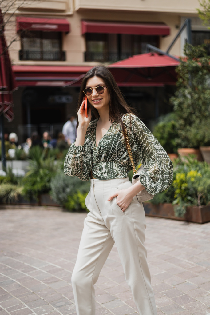 Весела молода жінка з довгим волоссям і сонцезахисними окулярами посміхається під час розмови на смартфоні і стоїть з рукою в кишені і сумочці на ланцюговому ремені біля розмитого будинку і рослин у Стамбулі.  - Фото, зображення