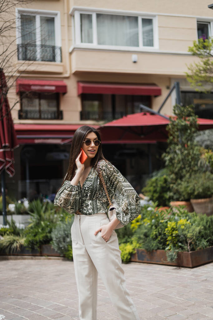 giovane donna con capelli lunghi e occhiali da sole sorridente mentre parla su smartphone e in piedi con mano in tasca e borsetta sul cinturino a catena vicino all'edificio sfocato e alle piante a Istanbul  - Foto, immagini