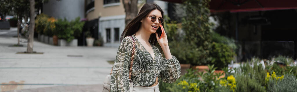 alegre joven con el pelo largo y gafas de sol sonriendo mientras habla en el teléfono inteligente y caminando con el bolso en la correa de cadena cerca del edificio borroso y plantas en la calle en Estambul, pancarta  - Foto, imagen
