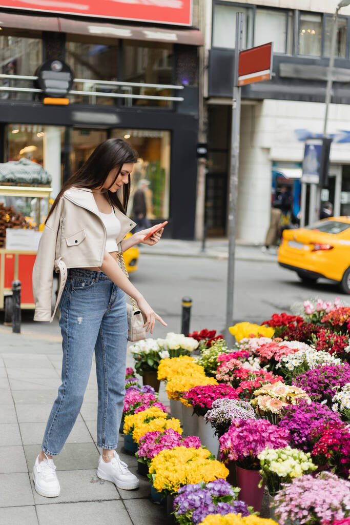 brunetka kobieta z długimi włosami stojąca w beżowej skórzanej kurtce i dżinsach jeansowych trzymając smartfona i wskazując na bukiety kwiatów obok zamazanego samochodu na ulicy w Stambule, sprzedawca - Zdjęcie, obraz