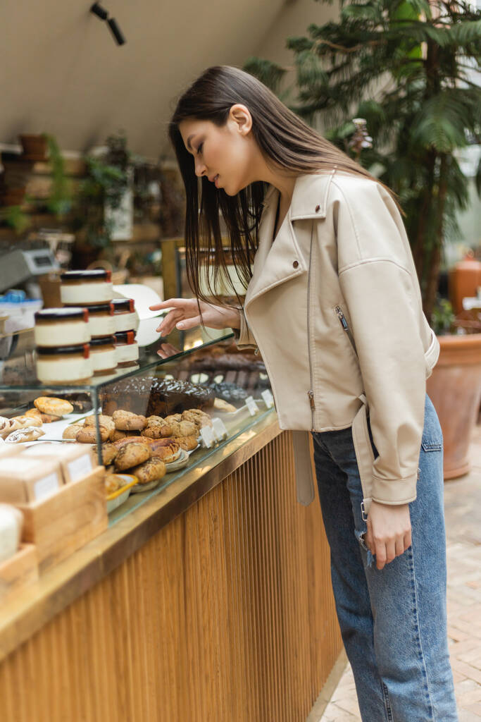 Jeune femme brune aux cheveux longs en veste en cuir beige et jean denim debout près de l'affichage du gâteau et choisissant la pâtisserie près des bocaux de confiture dans la boulangerie moderne à Istanbul  - Photo, image