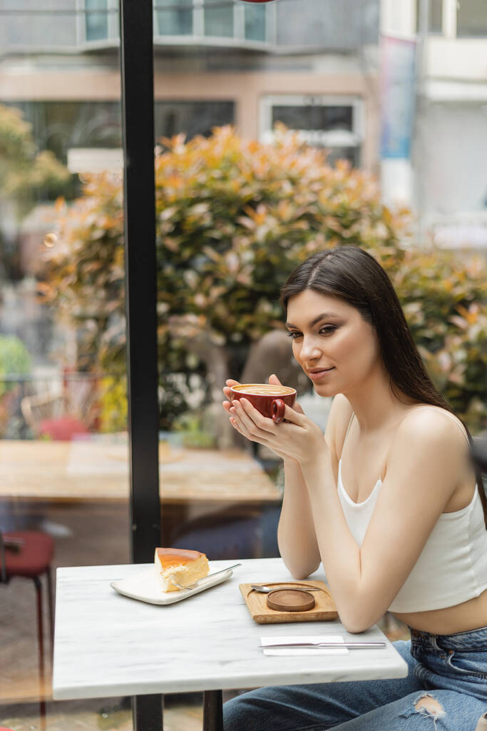 donna felice con i capelli lunghi seduta accanto alla finestra e che tiene una tazza di cappuccino con arte del caffè vicino cheesecake sul piatto del tavolo da bistrot all'interno del moderno caffè di Istanbul  - Foto, immagini