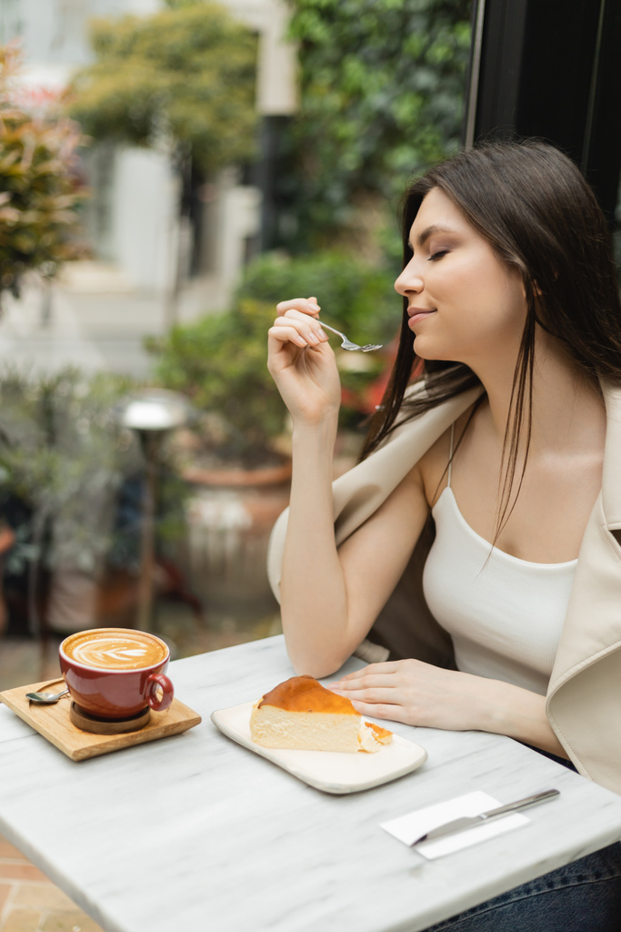 jovem com garfo de cabelo comprido perto de cheesecake ao lado da xícara de cappuccino na mesa de bistrô enquanto sentado em jaqueta de couro perto da janela dentro do café moderno em Istambul  - Foto, Imagem