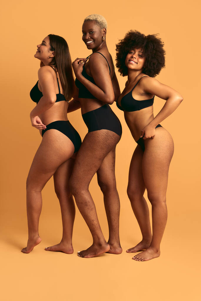 Vista laterale del corpo pieno di allegri giovani modelli femminili multietnici in biancheria intima nera in piedi contro lo sfondo giallo e sorridente felicemente in studio - Foto, immagini