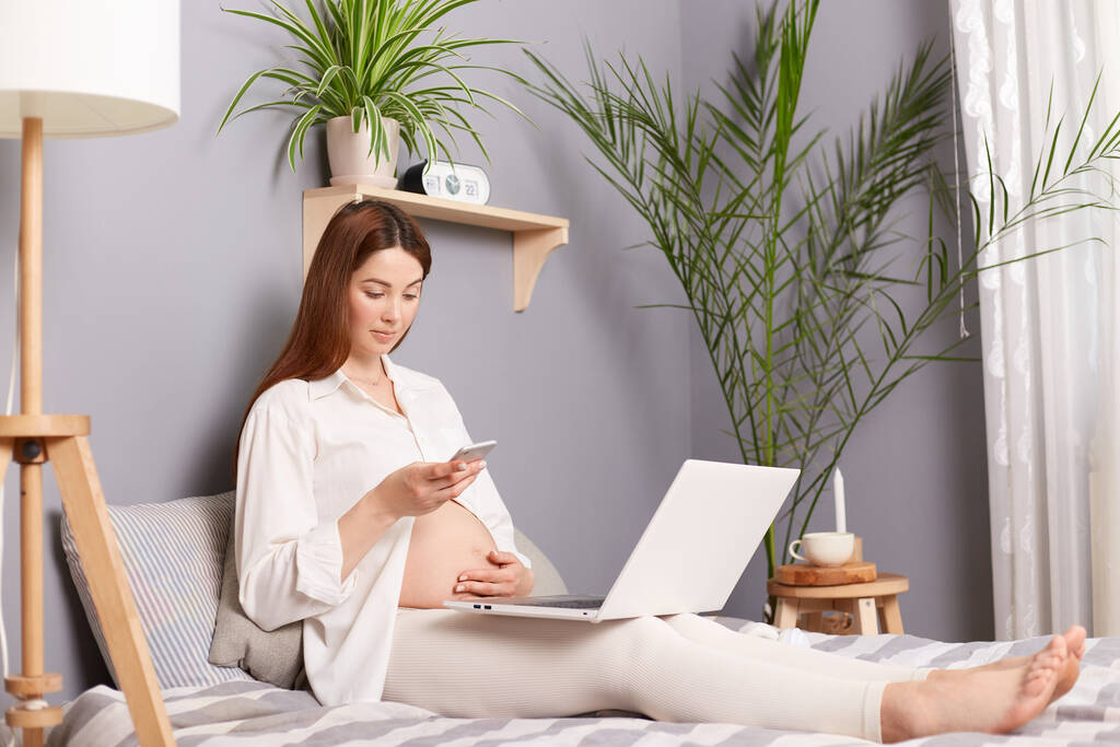 Kumral hamile bir kadın yatak odasında beyaz bir gömlek giyiyor bilgisayarında dinleniyor ve telefonuna bakıyor, konsantre bir ifadeye sahip, çıplak karnına dokunuyor.. - Fotoğraf, Görsel