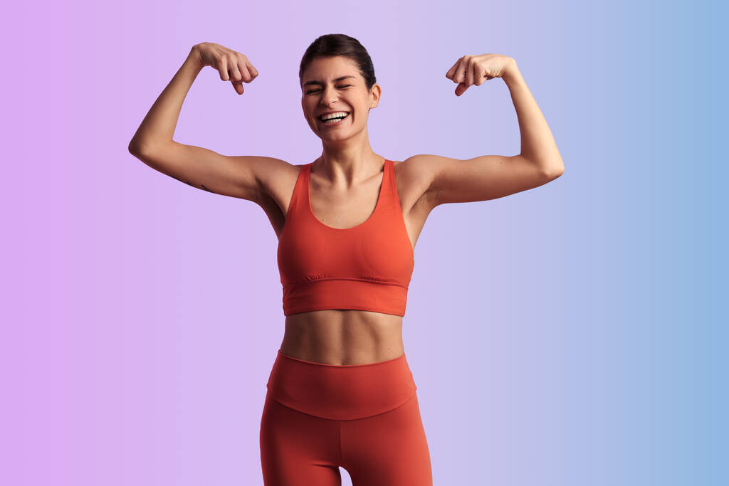 Giovane atleta donna in forma in abbigliamento sportivo in piedi con le braccia sollevate e mostrando bicipiti su sfondo sfumato - Foto, immagini