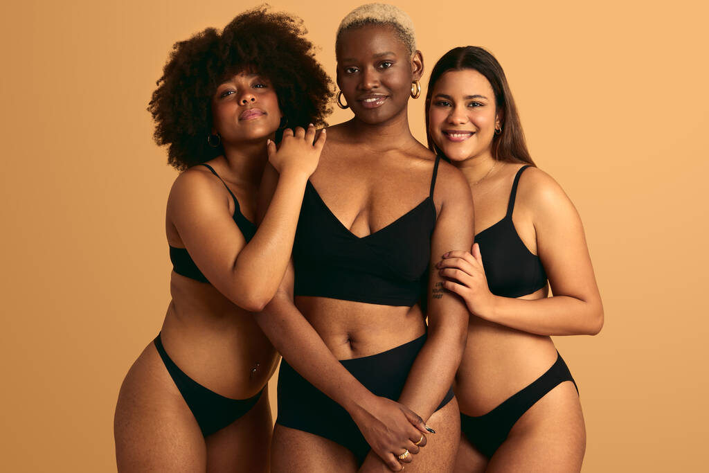 Fröhliche junge multiethnische Models in schwarzer Unterwäsche lächeln in die Kamera, während sie im Studio dicht an dicht vor beigem Hintergrund stehen - Foto, Bild