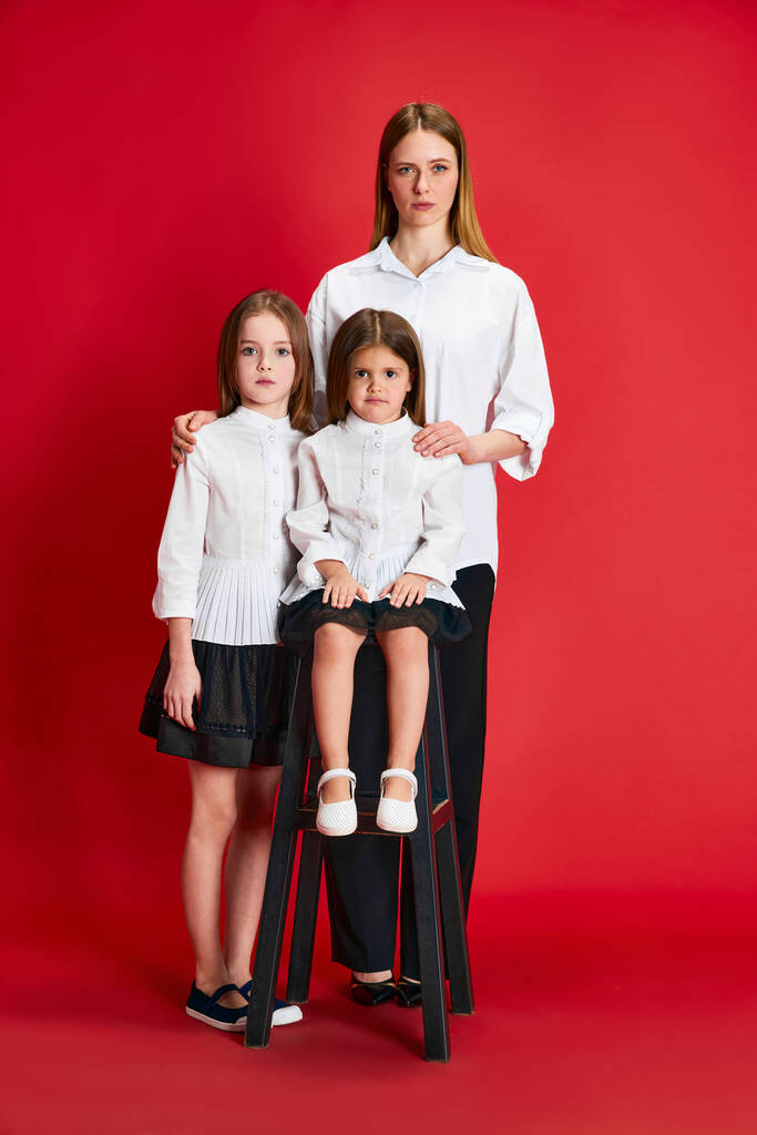Porträt der schönen Familie, Mutter und zwei kleine Mädchen, die in gleicher Kleidung vor rotem Studiohintergrund posieren. Konzept von Familie, Mutterschaft, Kindheit, Mode, Lebensstil, Liebe - Foto, Bild
