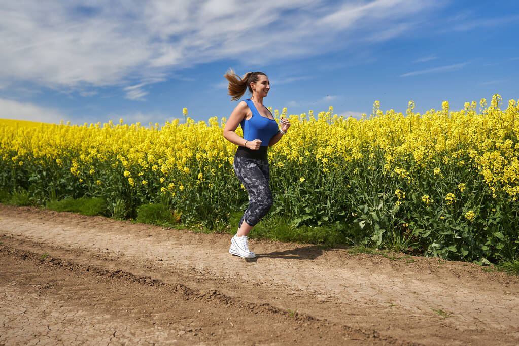 Plus size piękne latynoski kobieta jogging na polnej drodze przez pole rzepaku - Zdjęcie, obraz
