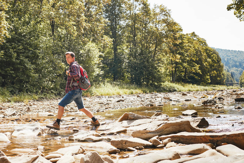 Trekking met rugzak concept imago. Backpacker draagt wandelschoenen die de bergrivier oversteken. Man wandelen in de bergen tijdens de zomer reis. Vakantie reis dicht bij de natuur. Natuurlijke omgeving - Foto, afbeelding